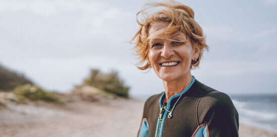 На фотографии изображена улыбающаяся женщина, наслаждающаяся моментом у моря, символизирующая спокойствие и радость, которые приносит пожизненное пенсионное страхование.