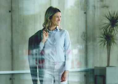 Женщина в светлой рубашке стоит у стеклянной стеной, держа пиджак на плече.