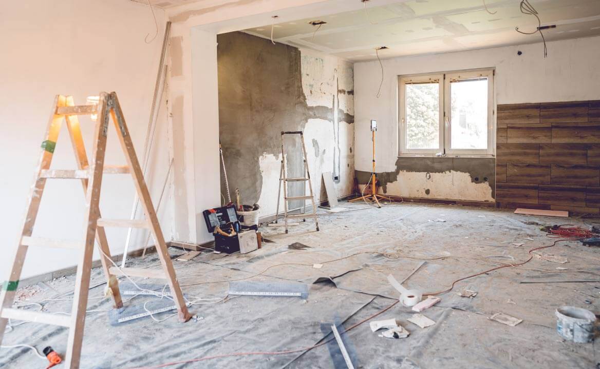 The room is under renovation – the walls have been freshly plastered and a protective sheet covers the floor. There are two construction ladders and toolboxes in the room. 