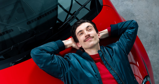 A man is resting on the hood of a red car, dressed in a blue jacket and a red shirt.