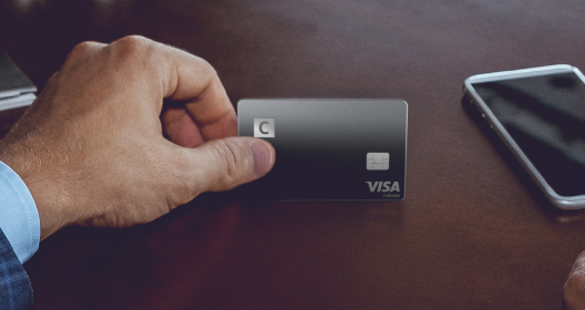 A man in a suit is holding a black Citadele C Infinite payment card with the C logo in the top left corner, focusing his attention on the card.