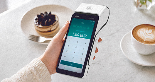 A hand holding a PAX payment terminal in a café, with a dessert plate and cappuccino in the background.