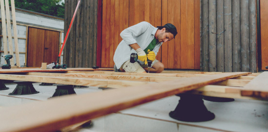 A man is building a new terrace using timber and is screwing in the boards with a drill. 