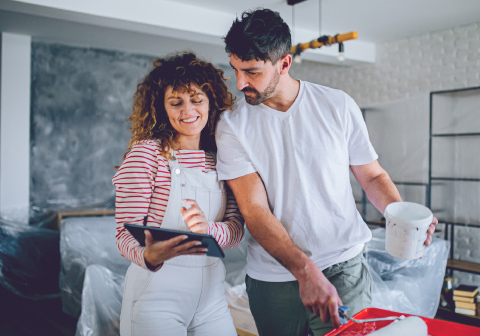 A woman is holding a tablet and showing a man what is happening on the screen. In the background, renovation work is taking place in the room; the man is holding a paint roller and a bucket. 