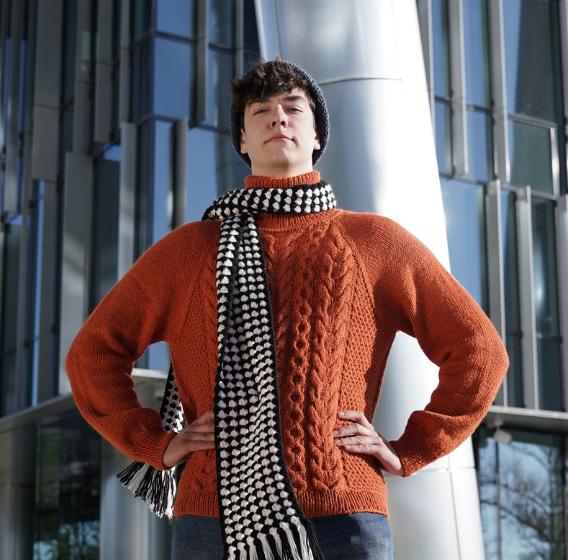 Человек в оранжевом свитере и шарфе стоит перед современной стеклянной фасадой здания.
