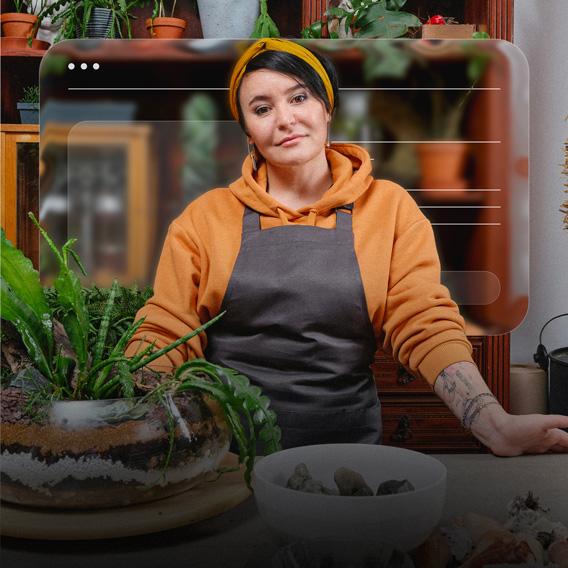 Человек в оранжевом свитере и сером фартуке стоит у прилавка с различными растениями.