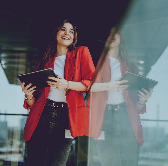 Женщина в красном пиджаке держит планшет у стеклянного фасада здания.