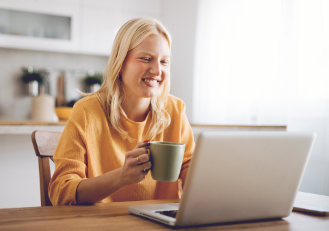A woman is sitting at a table with a cup of coffee in her hands, looking at a laptop. 