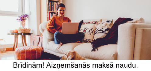 Sieviete sēž uz gaiša dīvāna ar klēpjdatoru, bet uz krāsaina paklāja priekšā guļ zelta retrīvers saulainā, mājīgā viesistabā.